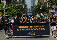 Members of the Audible community walk proudly in the 2022 New York City Pride Parade, holding a banner that says "Queer Stories by Queer Voices... Audible Pride" and holding rainbow-colored fans and flags.