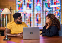 Two employees sit at a table in Audible's Innovation Cathedral in Newark. A laptop is open between them and behind them, slightly out of focus, is a wall of stained glass. A lamp sits on the table. They are looking at each other and smiling as they talk.