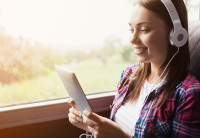 A girl sits by the window on a bus wearing a white t-shirt and a red and blue flannel shirt with silver headphones on. She's listening to a Audible book playig on the tablet she's holding. 