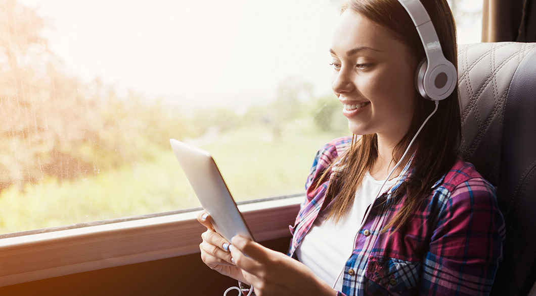A girl sits by the window on a bus wearing a white t-shirt and a red and blue flannel shirt with silver headphones on. She's listening to a Audible book playig on the tablet she's holding. 