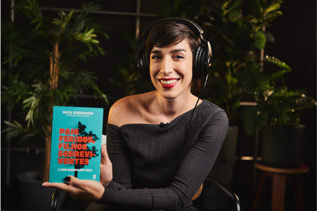 Woman in headphones smiling at camera, holding an audiobook titled "Pais Feridos Filhos Resilientes" in a decorated room with plants.