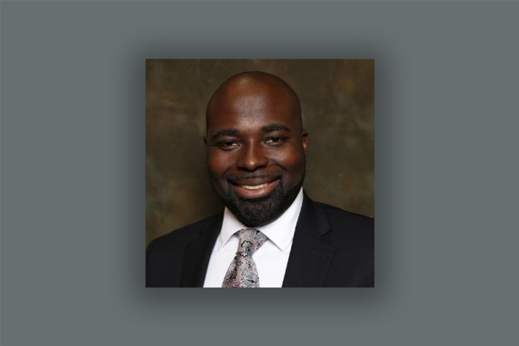 Audible employee St Jules Desir appears against a gray background wearing a blue suit, white button down shirt and pink and silver patterned tie. He is looking directly at the camera and smiling.