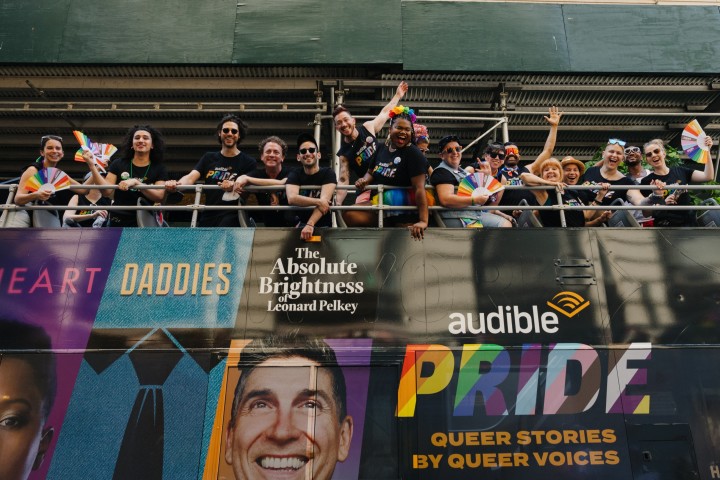 Audible community members cheer on top of a double decker bus. The bus is wrapped with Audible Originals.