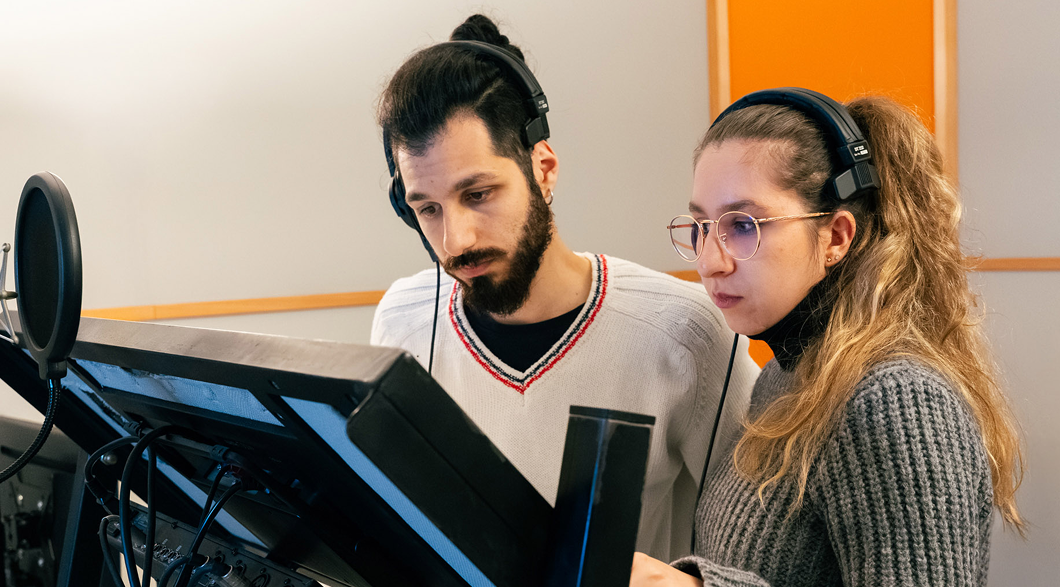 Two young actors stand with headphones on in front of a mic and stand holding a book they're narrating. 