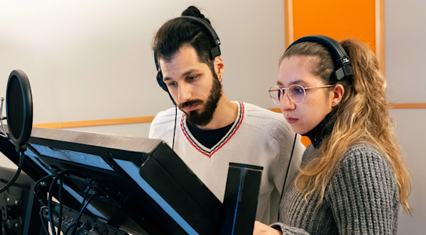 Two young actors stand with headphones on in front of a mic and stand holding a book they're narrating. 