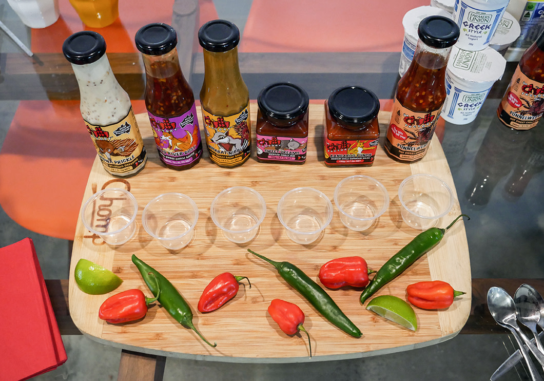 Several hot sauces and chilis line a cutting board with tasting cups in the middle.