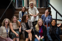 A group of Audible employees who are members and leaders of Women@Audible sit on the steps in our Innovation Cathedral building. Posed for a group photo, someone has obviously said something funny that has them all laughing and leaning into or looking at each other.  