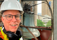 Mike Stevens smiles at the camera wearing a construction hard hat, in front of the historic steeple bell at Audible's Innovation Cathedral.