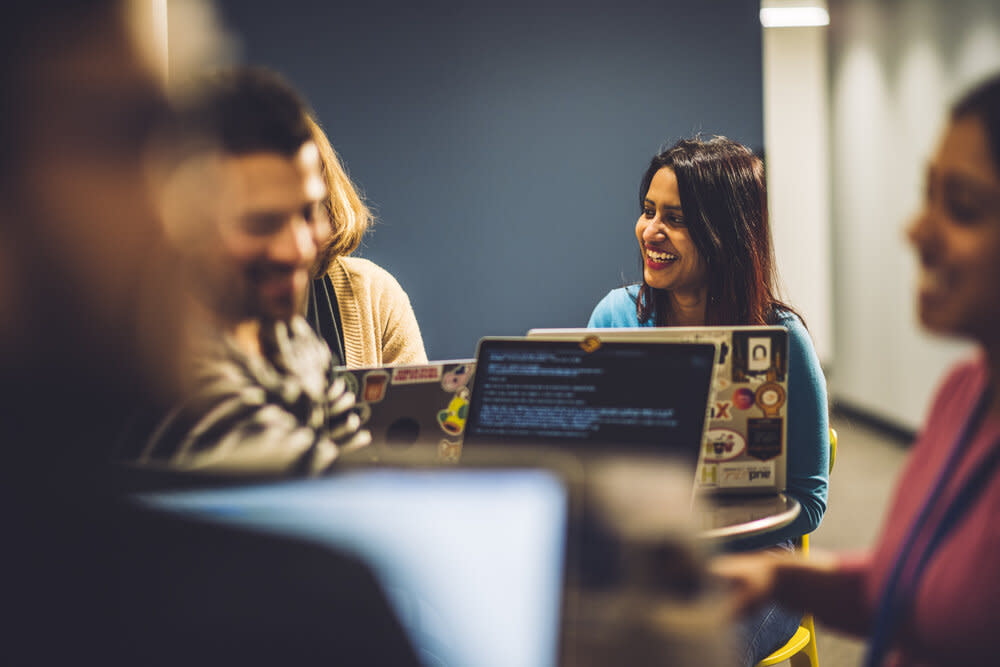 A group of Audible employees are gathered around a table in Audible's office. One woman smiles brightly to  her colleague sitting next to her.