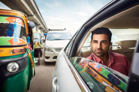 A man sits in the back of a car stuck in traffic on a road in India. His window is down and he has headphones on.