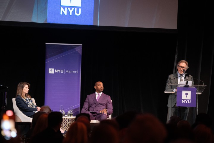 An event with two individuals seated on the stage alongside a lectern with the NYU logo where a third person is standing and speaking, in front of an audience in a dimly lit room. A screen displaying the NYU logo is visible in the background, with the words "NYU Alumni" on a banner beside the stage.