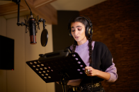 Actress Alba Flores stands in a studio recording the audio memoir "Mujer de Frontera" for Audible Spain. She has her hands holding the sides of the stand in front of her and a large microphone hangs down before her. She has headphones on and is wearing a purple and black sweater.  