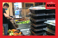 Boxes of meals sit on a kitchen counter in a restaurant waiting to be delivered to Newark residents in need. Beyond them a restaurant workers is filling an empty container with chicken and greens to make up another meal for delivery.