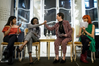 Four women sit on stage talking on a panel. The middle two women are clasping hands in a type of high five.