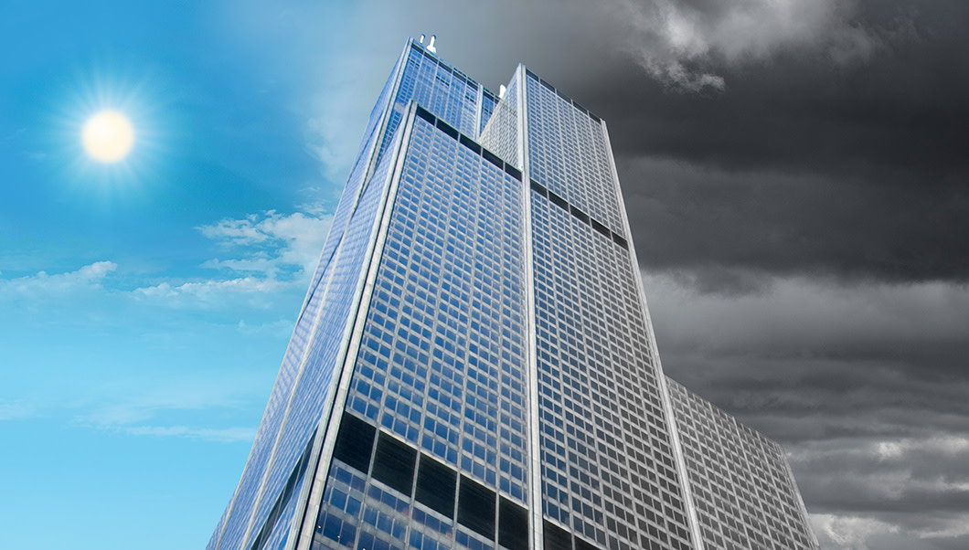 Staring up the side of a tall glass office building with blue sky on one side of it and dark clouds on the other.