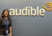 Audible employee Nandhini Gunalan, who participated in the 'New Chapter' Returnship Program, stands in our Newark office by an Audible logo on the wall. She is wearing a black shirt and her ID and lanyard and smiling. 