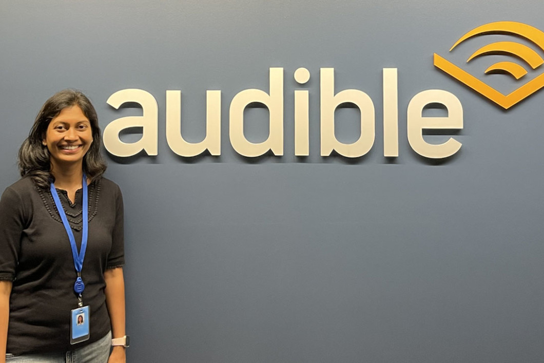 Audible employee Nandhini Gunalan, who participated in the 'New Chapter' Returnship Program, stands in our Newark office by an Audible logo on the wall. She is wearing a black shirt and her ID and lanyard and smiling. 