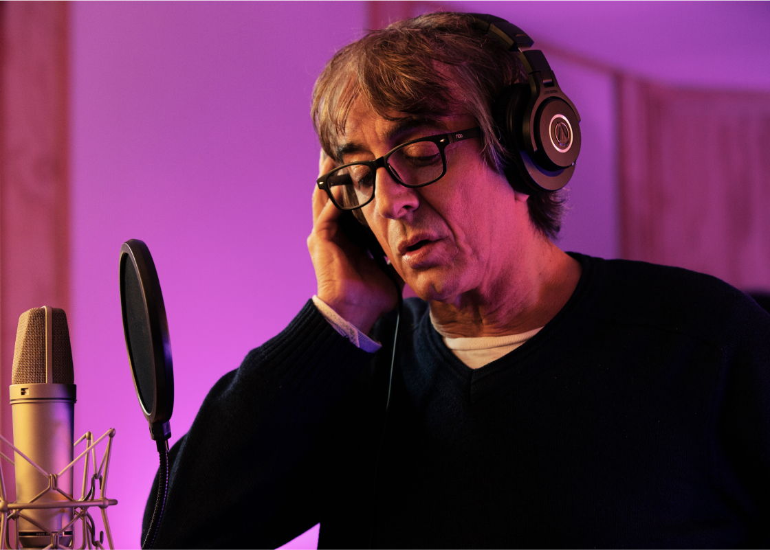 Luca Ward stands at a microphone in an Audible studio performing an audiobook. There are black headphones over his ears.