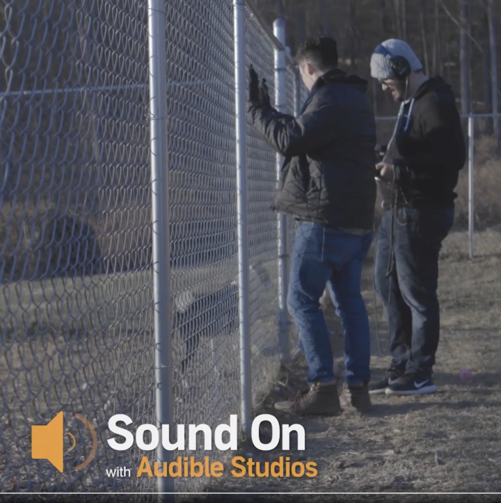 Two Audible sound designers stand by a wire fence at a dog shelter. One has his hands on the fence, the other is wearing headphones over a warm winter cap while holding a microphone to record the dog on the other side of the fence. They are wearing warm clothes--gloves, parka, hoodie, jeans. In the bottom of the picture there is a volume symbol and the words "Sound On with Audible Studios".