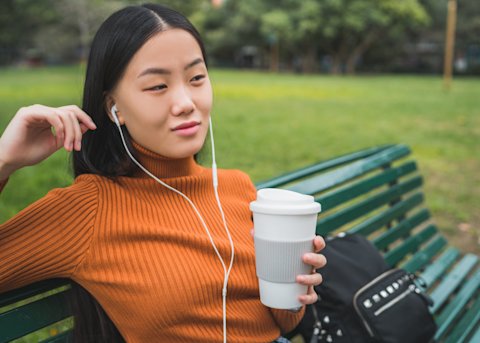 A woman sits on a park bench listening to an audiobook through the headphones in her eats while holding a coffee cup. Her bag sits on the bench beside her.