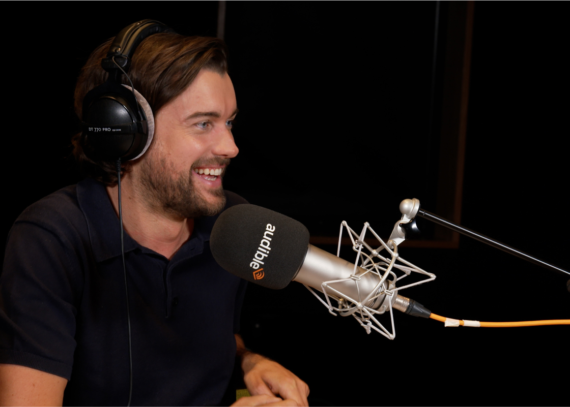 Jack Whitehall sits in front of a microphone in an Audible recording studio, black headphones over his ears. 