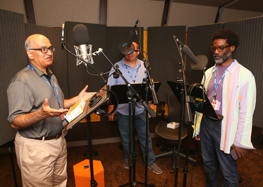 Three men stand at large microphones and music stands in front of them that hold scripts in an audio studio. Black noise absorbent material is behind them. Avid listeners, they are getting a chance to see what it's like to narrate an audiobook for Audible as part of a member appreciation day.