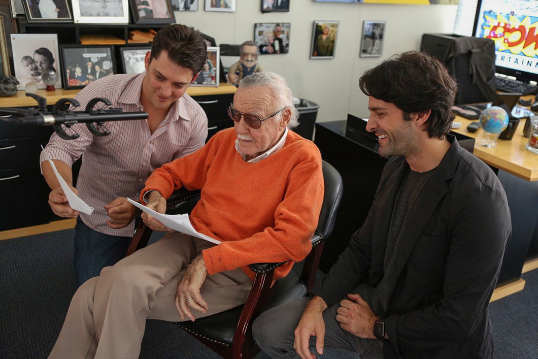 Stan Lee sits in front of a microphone reviewing text with two collaborators