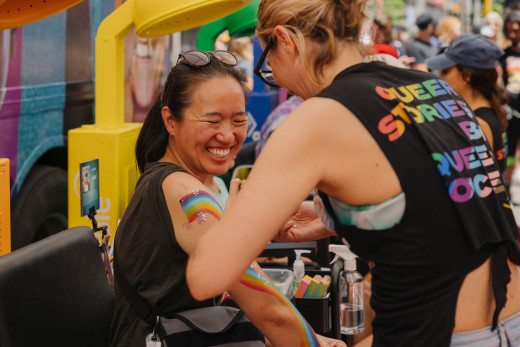 A woman smiles in joy while getting a rainbow painting on her arm. The artist has her back facing the camera, and you can read "Queer Stories by Queer Voices" on the back of her shirt.