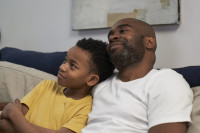 A father and son sit on the couch together. The child is leaning into his dad's shoulder and the dad has his arm wrapped around his son. The look happy and relaxed as they sit there listening together.
