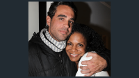 Actor Bobby Cannavale stands with his arm around actor Audra McDonald, holding her close. She is smiling and they are both looking directly into the camera. He is wearing a black zip up jacket with a gray and white scarf wrapped around his neck. She has on a white long sleeve shirt.