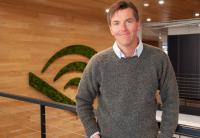 Matthew Thornton is standing in the Audible ofices looking directly at the camera. Behind him is a wooden wall with the Audible logo created in greenery. 