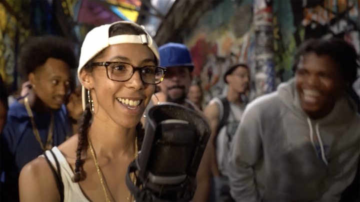 In a screenshot from the video shared as part of this post, a girl stands in front of a large microphone, wearing a white baseball cap on backwards and glasses. There is a group of people behind her, mostly men, listening and getting into what she is putting down.