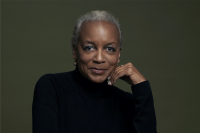 Yvonne Durant sits looking straight to camera in a photo shot against a gray background. She is wearing a dark turtleneck and has short silver hair, an upturned smile, and her face is resting ever so slightly against her hand.   