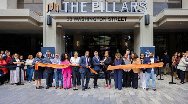 A crowd is gathered in front of a building called "The Pillars" in downtown Newark with people cutting a large orange ribbon to mark its opening.