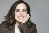 Rachel Ghiazza, Audible EVP, Head of US Content, is looking straight at the camera in a portrait shot set against a gray background. Her head and shoulders can be seen. She is smiling, wearing a brown tweed blazer, and her face is resting in her hand.
