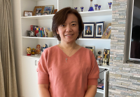 Japan Country Manager Shima Osaka stands in the living room of her home. Behind her, bookshelves are filled with family photos, blue glass, books and art. She is smiling directly at the camera and is wearing a peach v-neck sweater.