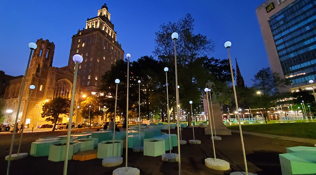 Solar lights and green hexagon planters and seats are artfully arranged in Washington Park with Audible's headquarter buildings lit up in the background.
