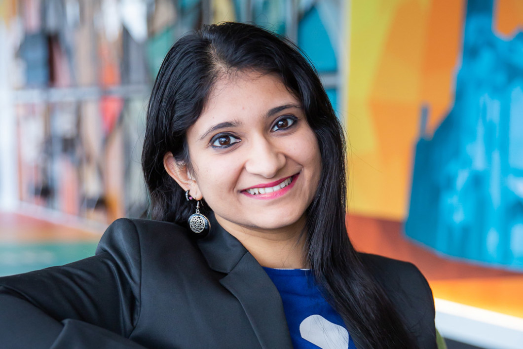 Audible employee Akanksha Gupta sits in Audible's Newark headquarters lobby wearing a blue shirt and grey blazer. She is looking straight at the camera and is smiling. Behind her is a multi-colored mural that is blurred to keep her the main central focus. 