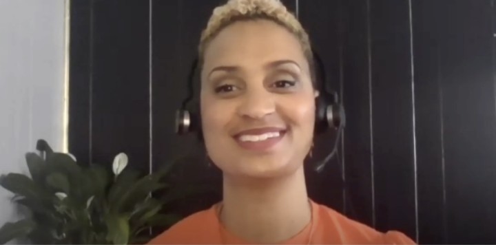 Audible VP, Urban Innovation, Aisha Glover sits talking on camera for the SXSW panel. She is wearing headphones and an orange sweater. There is a flowering plant in the background.