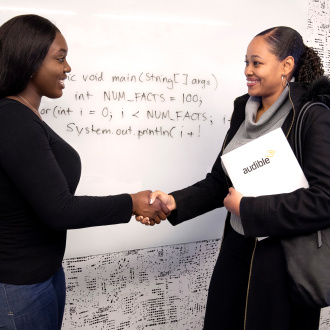 Two women stand shaking hands in front of a whiteboard as if at the end of a job interview. One holds a three ring binder with Audible written across the front.