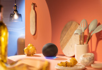 A black Amazon Echo device is show in the middle of a kitchen workspace among cutting boards, bowls, and wooden spoons.