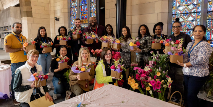 A large group of Audible employees are posing in a group with floral arrangements from a team building event. They are standing in front of a stained glass window in the Innovation Cathedral.
