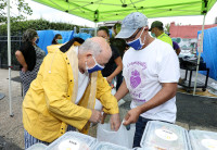 A community volunteer holds a bag filled with Newark Working Kitchens meals for an elderly Newark resident who is reaching into the bag. They are in a pop up distribution area, with tents and tables set up in an urban area.