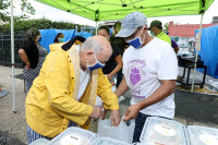 A community volunteer holds a bag filled with Newark Working Kitchens meals for an elderly Newark resident who is reaching into the bag. They are in a pop up distribution area, with tents and tables set up in an urban area.