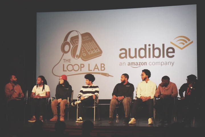 Students at Loop Lab sit on a stage with the Loop Lab and Audible logos in the background.