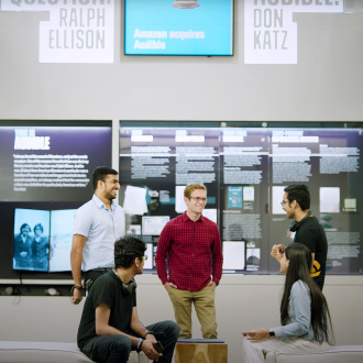 A group of tech interns gathers in a group of the lobby of our Innovation Cathedral in downtown Newark where a lot of our tech teams work. Information about the history of Audible are on the wall behind them. They are all smiling and laughing.