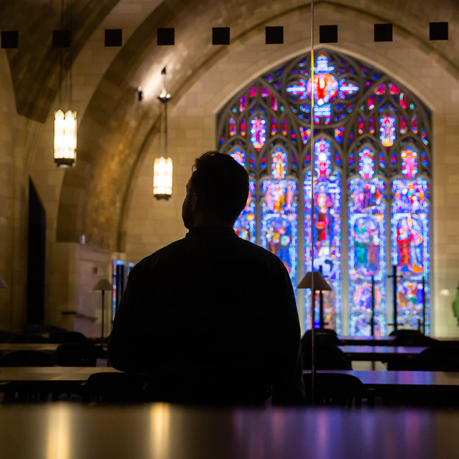 A man sits at a table in a library like room. We see him from behind against the light of a stained glass window.