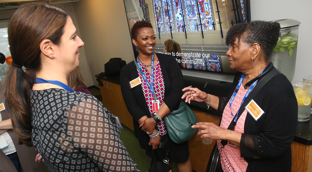Three women stand talking in a group. They're smiling and animated. In the background is a bit of a staircase and stained glass wall from Audible's Innovation Cathedral, a restored church that now operates as a office.