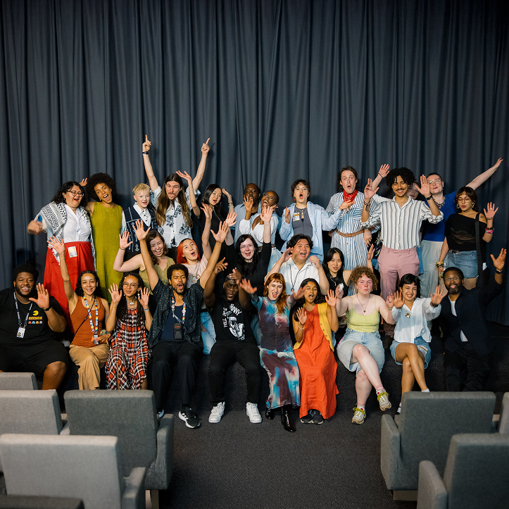 A group of young actors three rows deep are collected on the lip of a stage, the curtain closed behind them, all raising their arms in excitement.