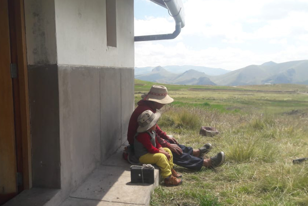 A father and son sit side by side leaning against a house on a sunny day looking out over a green field in Peru. A radio sits beside them. In the distance, mountains rise on the horizon.